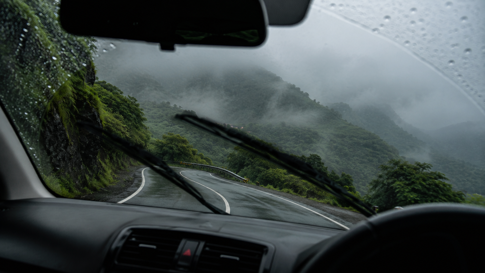 rain-speckled windshield at a winding ghat road disappearing into fog-covered hills, wipers mid-motion
