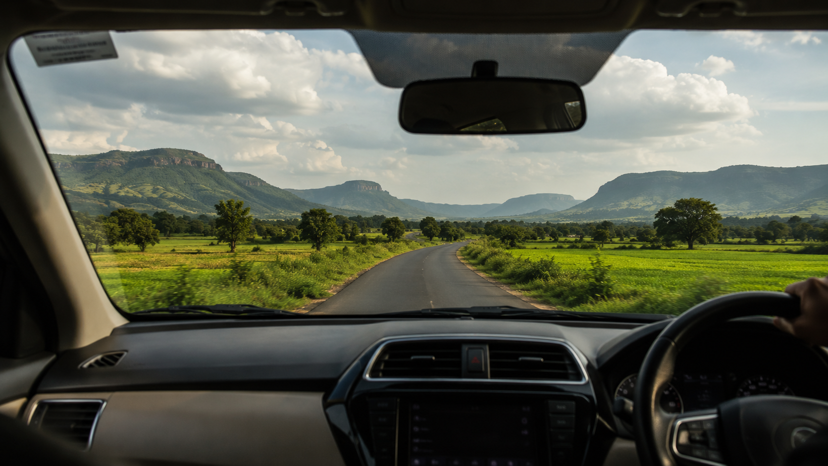 View through a car windshield of the narrow village road leading into Bhandardara surrounded by green fields