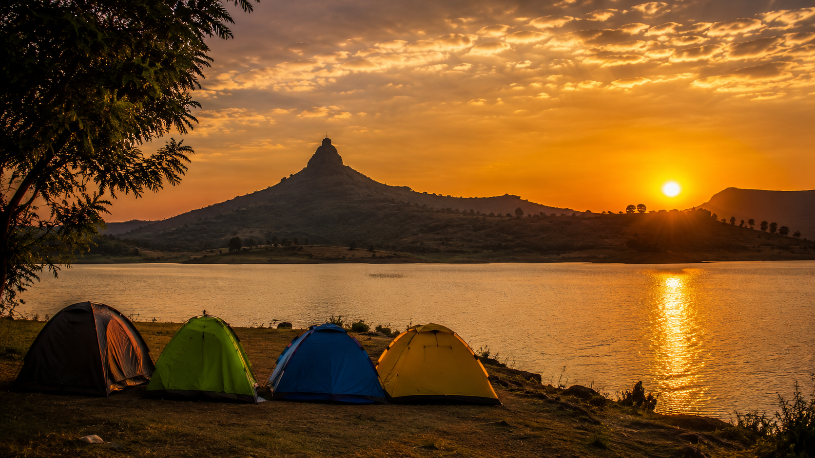 Tents pitched beside Pawna Lake at sunset with Tikona Fort silhouetted on the horizon